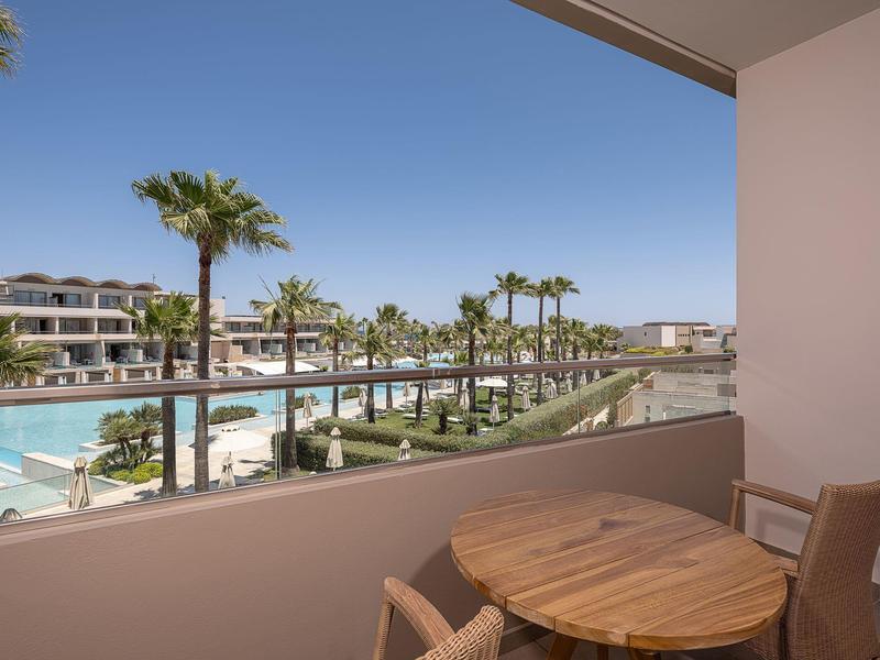 Vista desde un balcón con mesa hacia una zona de piscina con palmeras y cielo claro.