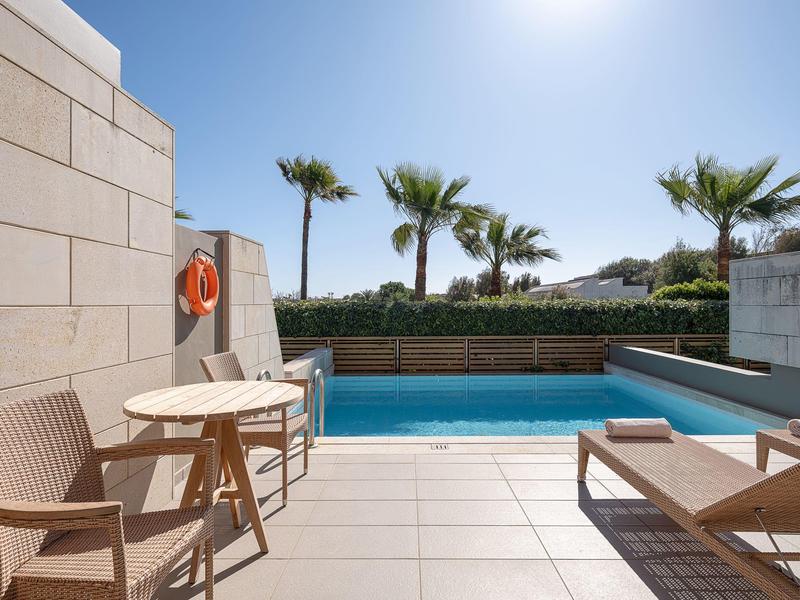 Zona de piscina con tumbonas, mesa y palmeras al fondo bajo cielo azul.