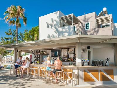 Área de bar al aire libre moderna con asientos y clientes, rodeada de palmeras y cielo azul.