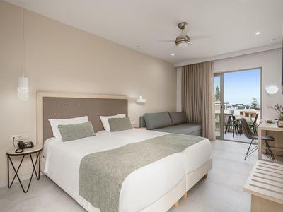 Habitación de hotel moderna con cama doble, sofá gris y balcón con vistas exteriores.