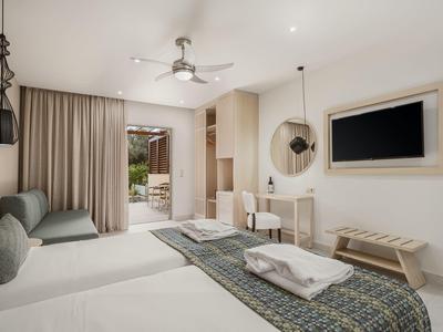 Habitación de hotel luminosa con dos camas, sofá, escritorio, TV y acceso a una terraza con vista verde.