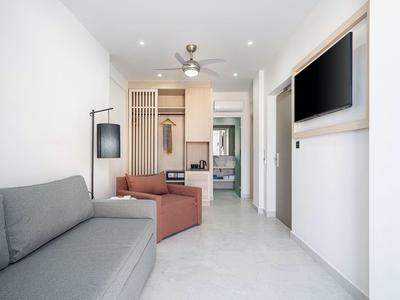 Sala de estar luminosa con sofá gris, sillón rojo, ventilador de techo y TV en la pared.