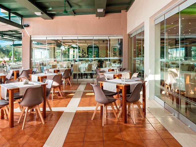 Terraza de hotel luminosa y moderna con mesas y sillas, rodeada de paredes de cristal y naturaleza verde.