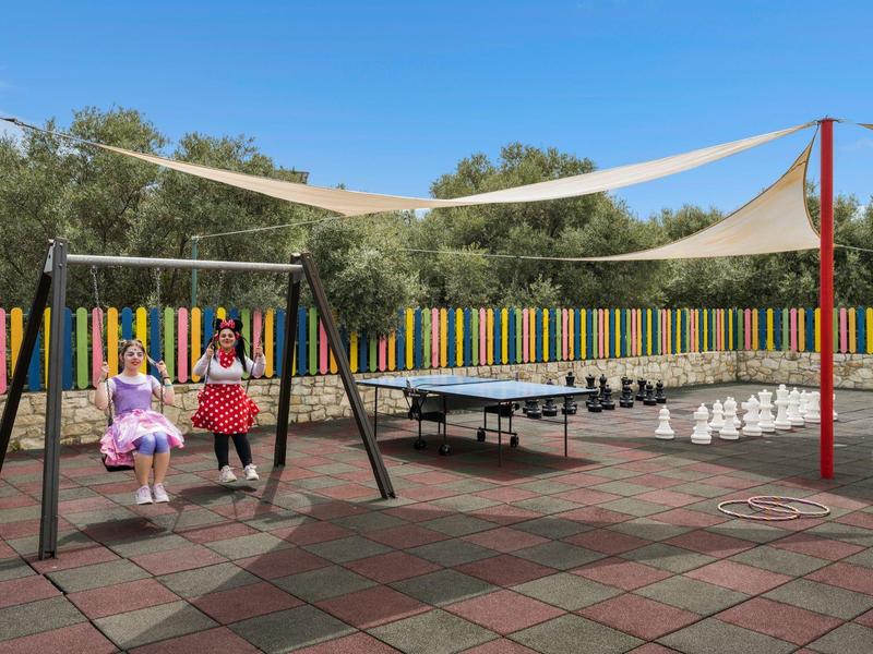 Área de juegos con dos niños en columpios, mesa de ping-pong y velas de sombra al aire libre.