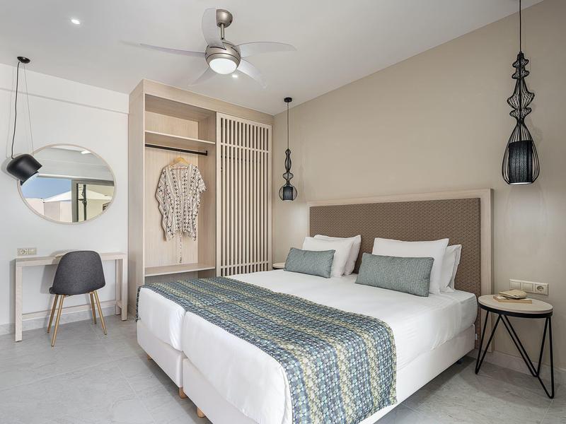 Habitación de hotel moderna con cama doble, escritorio y pequeño armario en tonos neutros.