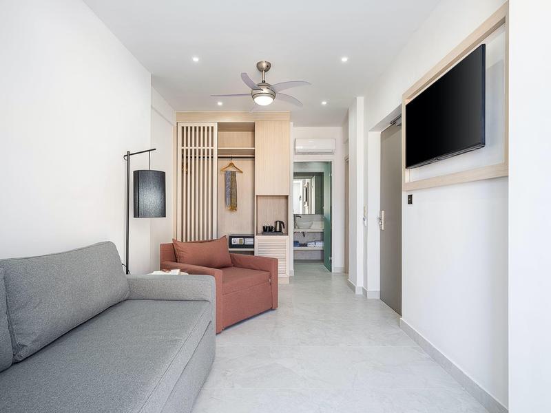 Sala de estar luminosa con sofá gris, sillón rojo, ventilador de techo y TV en la pared.