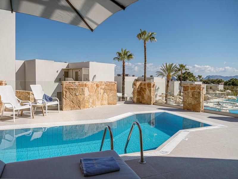 Piscina al aire libre moderna con tumbonas y sombrilla, rodeada de palmeras y montañas.