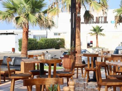 Gemütlicher Außenbereich eines Restaurants mit Holztischen, Stühlen und Palmen bei sonnigem Wetter.