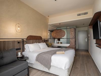 Nowoczesny pokój hotelowy z dużym łóżkiem, szarą sofą, drewnianą podłogą i telewizorem na ścianie.