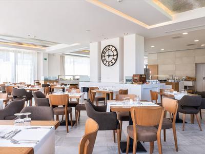 Jasna restauracja z nowoczesnymi drewnianymi stołami i krzesłami, duży zegar na ścianie.