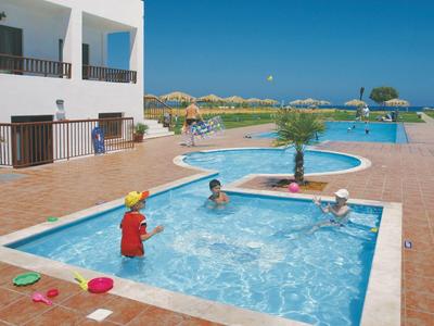 Dzieci bawią się w płytkim basenie obok morza i tarasu z białymi budynkami.