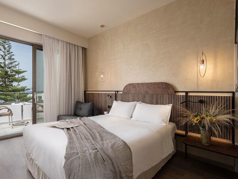 Jasny pokój hotelowy z łóżkiem queen, szarym kocem, dwoma fotelami, balkonem i beżowymi ścianami.