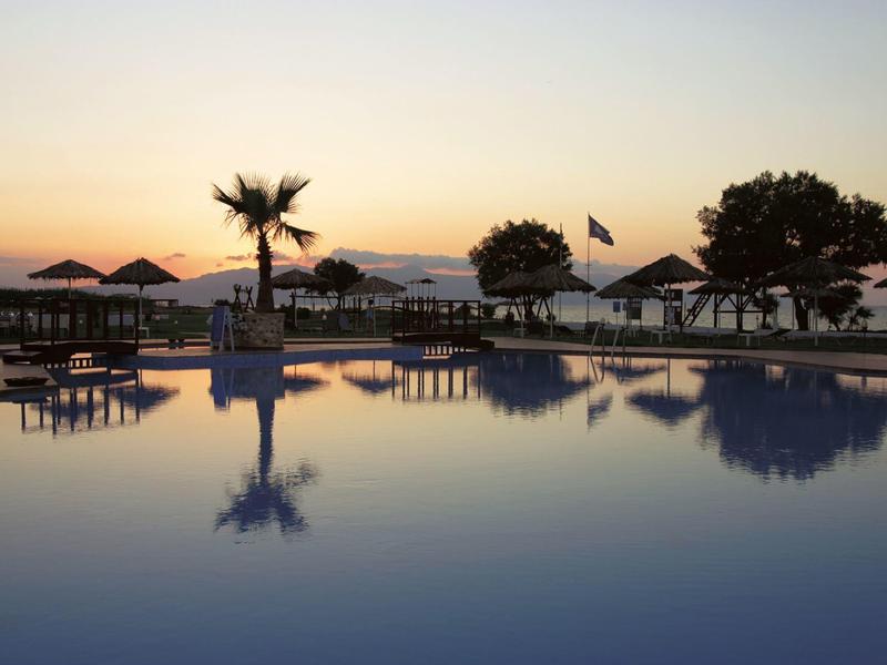 Basen o zachodzie słońca z palmami i parasolami odbijającymi się w wodzie.