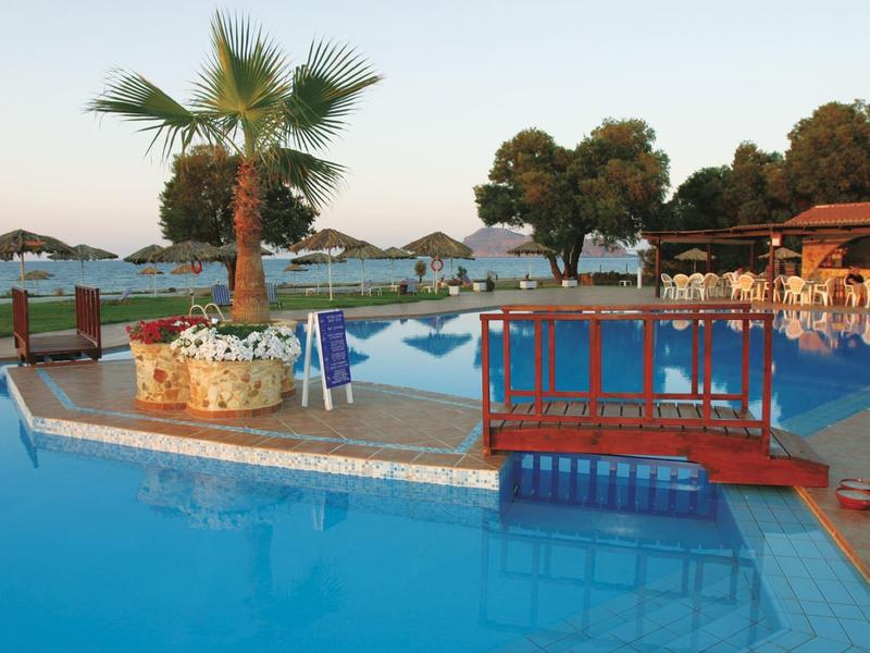 Strefa basenowa z mostkiem, palmami, parasolami i leżakami przy wodzie.