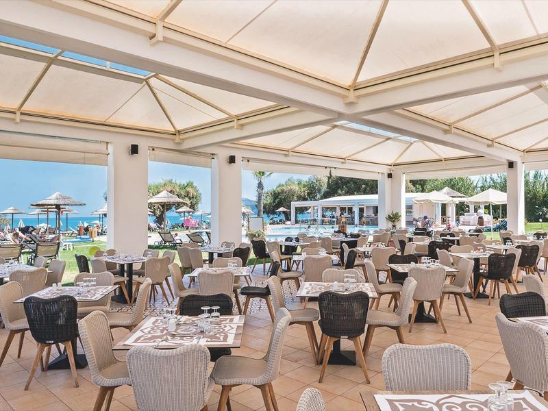 Restauracja na świeżym powietrzu z nakrytymi stołami, widokiem na plażę i morze z parasolami.