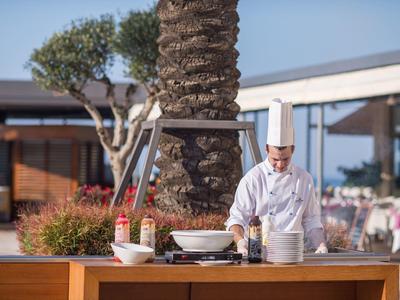 Chef in white uniform preparing food outdoors at a hotel with palm trees around.