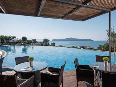 Venkovní část hotelu s bazénem a výhledem na moře, stoly a židle pod dřevěným přístřeškem.