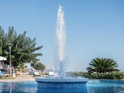 Piscine d'hôtel moderne avec fontaine centrale et parasols à côté de plantes vertes sous ciel clair.