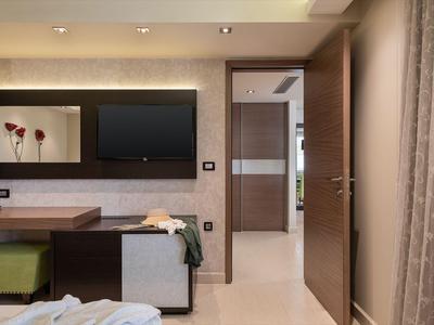 Espace moderne de chambre d'hôtel avec télévision, bureau, chaise et portes ouvertes vers un couloir lumineux.