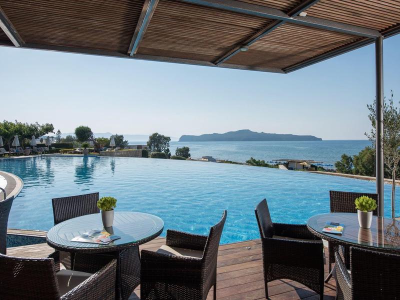 View of a pool with tables and chairs under a wooden canopy with sea and island in the background