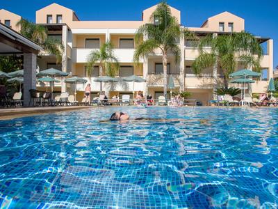 Hotelgebäude mit mehreren Stockwerken, Pool im Vordergrund und Palmen auf einer sonnigen Terrasse.