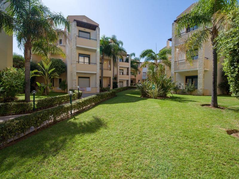 Green lawn area between two apartment buildings with palm trees and clear blue sky.