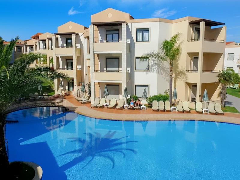 Modern hotel complex with large pool, palm trees, and sun loungers under clear sky.