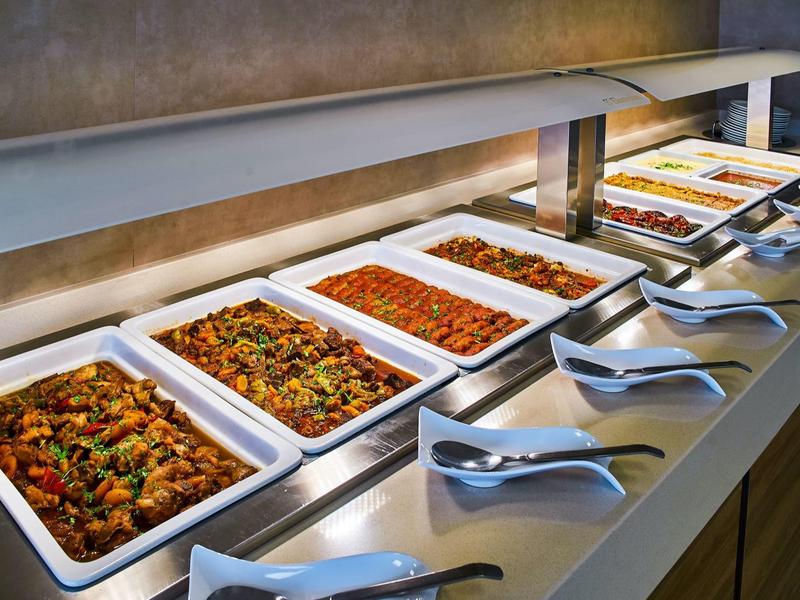 Buffet with various hot dishes in white containers and serving spoons.