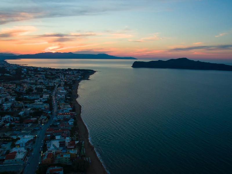 Aerial view of a coastal town at sunset with calm sea and island on the horizon.
