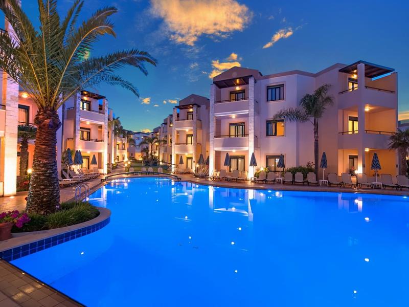 Illuminated pool area in front of a hotel at dusk with palm trees.