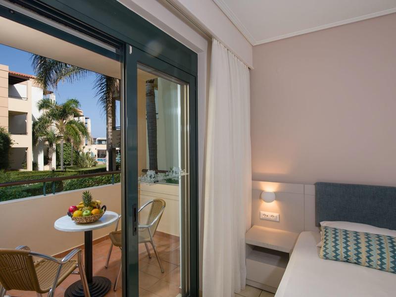 Hotel room with double bed and view of balcony with table and chairs, garden with palm trees beyond.