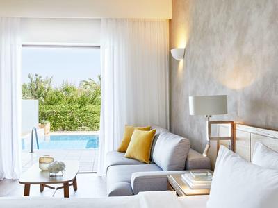 Moderní hotelový pokoj s pohovkou, polštáři a výhledem na bazén přes otevřené skleněné dveře.