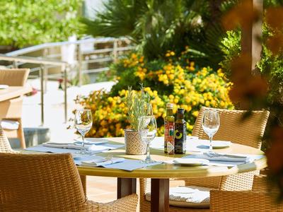 Venkovní stoly restaurace s bambusovými židlemi, stolované s vinnými sklenicemi a jídelními lístky, obklopené rostlinami.