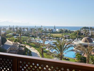 Blick von Balkon auf Hotelanlage mit Pools, Palmen und Meer im Hintergrund.