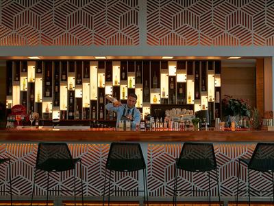 Moderní bar s geometrickými vzory na pultu a vysokých židlích, osvětlené police s lahvemi.