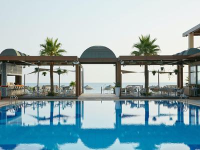 Hotelpool mit Liegestühlen und Meerblick, umgeben von Palmen und moderner Architektur.