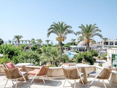 Terrasse avec chaises, vue sur des palmiers, piscine et bâtiments de l'hôtel par une journée ensoleillée.