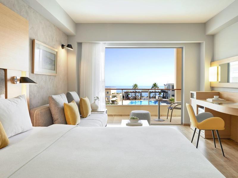 Světý hotelový pokoj se dvěma postelemi, posezením, psacím stolem a balkónem s výhledem na moře.