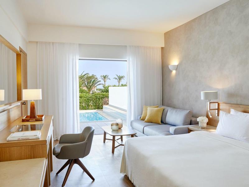 Světle hotelový pokoj s manželskou postelí, posezením a výhledem na bazén a palmy.