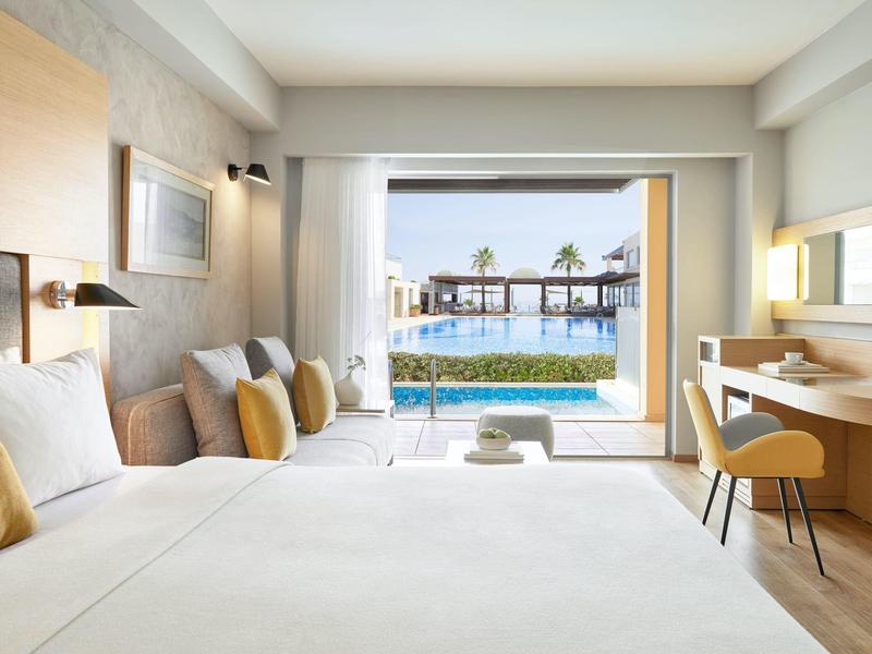 Světly hotelový pokoj s velkou postelí, posezením, stolem a výhledem na bazén přes skleněné dveře