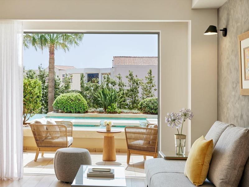 Moderní obývací prostor s pohovkou, křesly a výhledem na zahradu s bazénem a palmami.