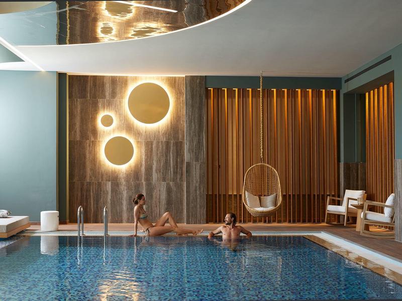 Vnitřní bazén s moderním kulatým osvětlením, dvě osoby ve vodě a závěsná židle.