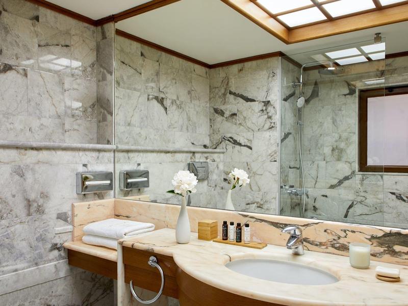 Moderní interiér koupelny s mramorovými zdmi, velkým zrcadlem a dřevěnou umyvadlovou skříňkou.