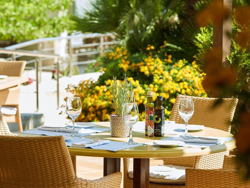 Venkovní stoly restaurace s bambusovými židlemi, stolované s vinnými sklenicemi a jídelními lístky, obklopené rostlinami.