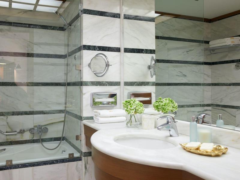 Moderní koupelna s mramorovými stěnami, skleněným sprchovým koutem a kulatým umyvadlem s dekoracemi.