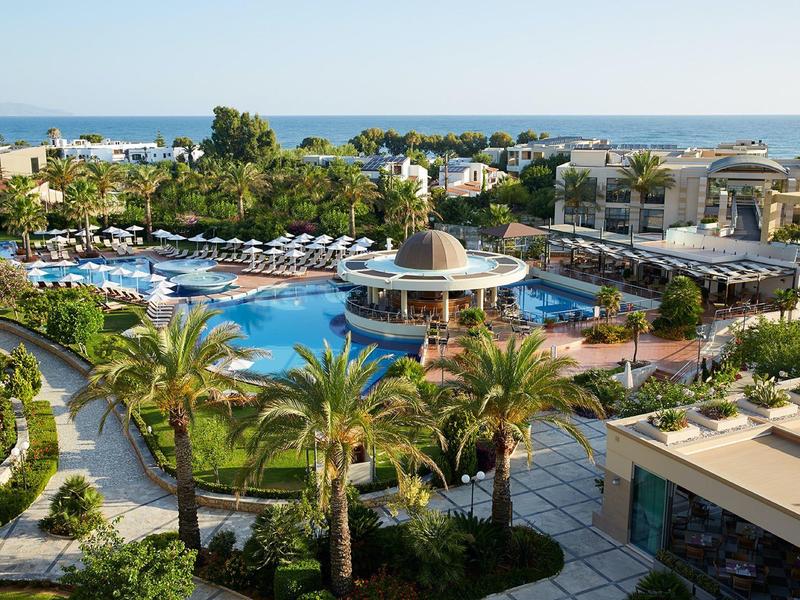 Rezort s velkým bazénem, palmami, lehátky a výhledem na moře v pozadí.