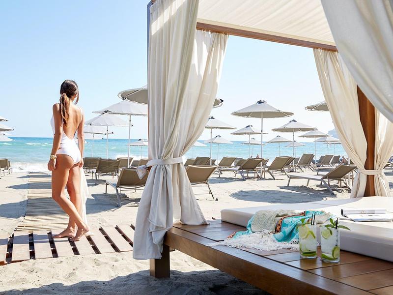 Žena kráčející po písečné pláži vedle zastíněného lehátka s nápoji pod baldachýnem