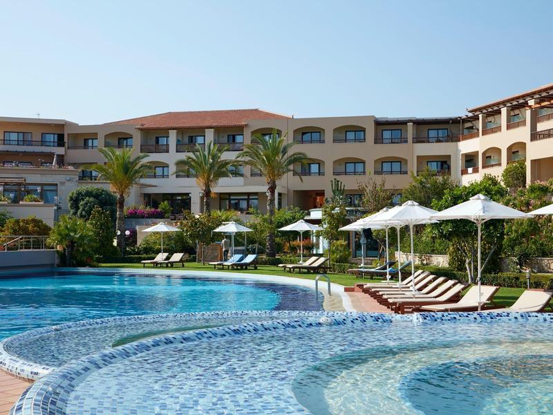 Hotel s velkým venkovním bazénem, palmami a lehátky pod bílými slunečníky.