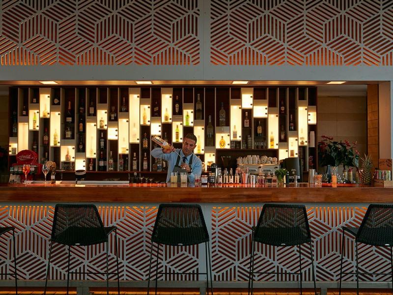 Moderní bar s geometrickými vzory na pultu a vysokých židlích, osvětlené police s lahvemi.