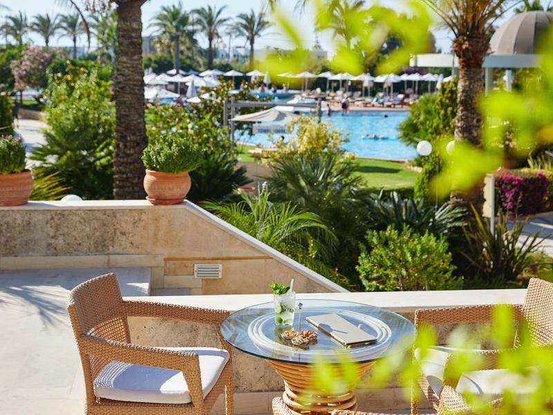 Venkovní terasa se dřevěnými židlemi a stolem s výhledem na bazén a palmy.